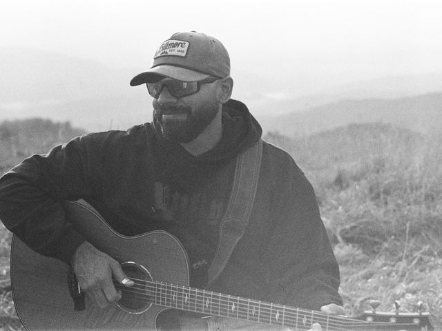 Black and white film photo of Josh Q smiling while playing guitar