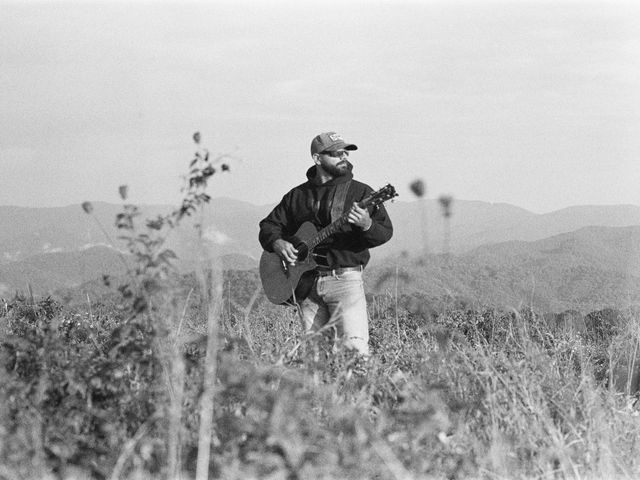 Black and white film photo of Josh Q standing with his guitar in a mountain field